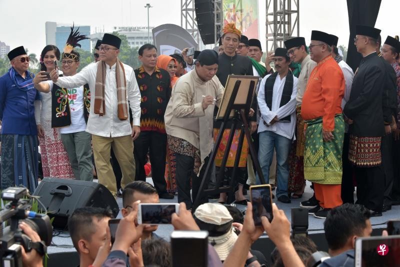 印尼總統佐科（右五）在儀式上望著其競選對手普拉博沃（中）握筆簽署和平競選宣言。各候選人穿著印尼各地的傳統服裝出席活動，佐科穿的是一件黑色的峇厘島民族服裝，而普拉博沃穿著一件褐色傳統中爪哇民族服裝。（圖源：AFP）