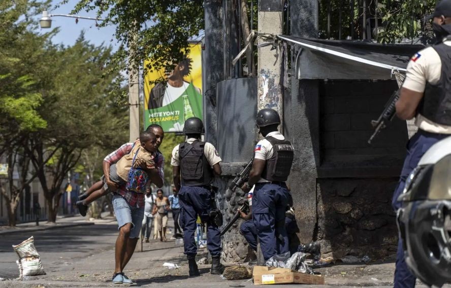 当地时间2023年3月3日，海地首都太子港总统府附近，一名男子抱着孩子经过海地国家警察，试图击退黑帮。（图：AFP）