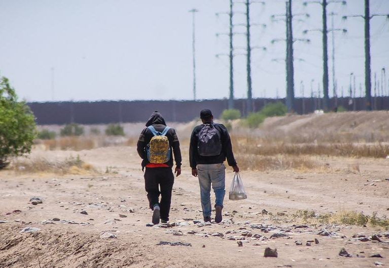 两名男子走在分隔墨西哥和美国的边境墙旁边。（图：国际移民组织）