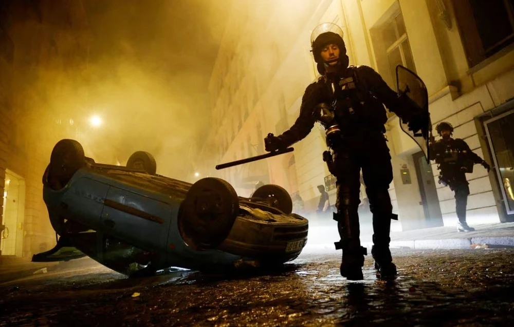 Các sĩ quan cảnh sát chống bạo động Pháp đi cạnh một chiếc xe bị lộn ngược ở Paris, Pháp vào ngày 2-7 - ngày thứ 5 của cuộc biểu tình sau cái chết của một thiếu niên 17 tuôi bị một sĩ quan cảnh sát Pháp bắn chết vì không tuân thủ hiệu lệnh của cảnh sát khi tham gia giao thông. Ảnh: REUTERS