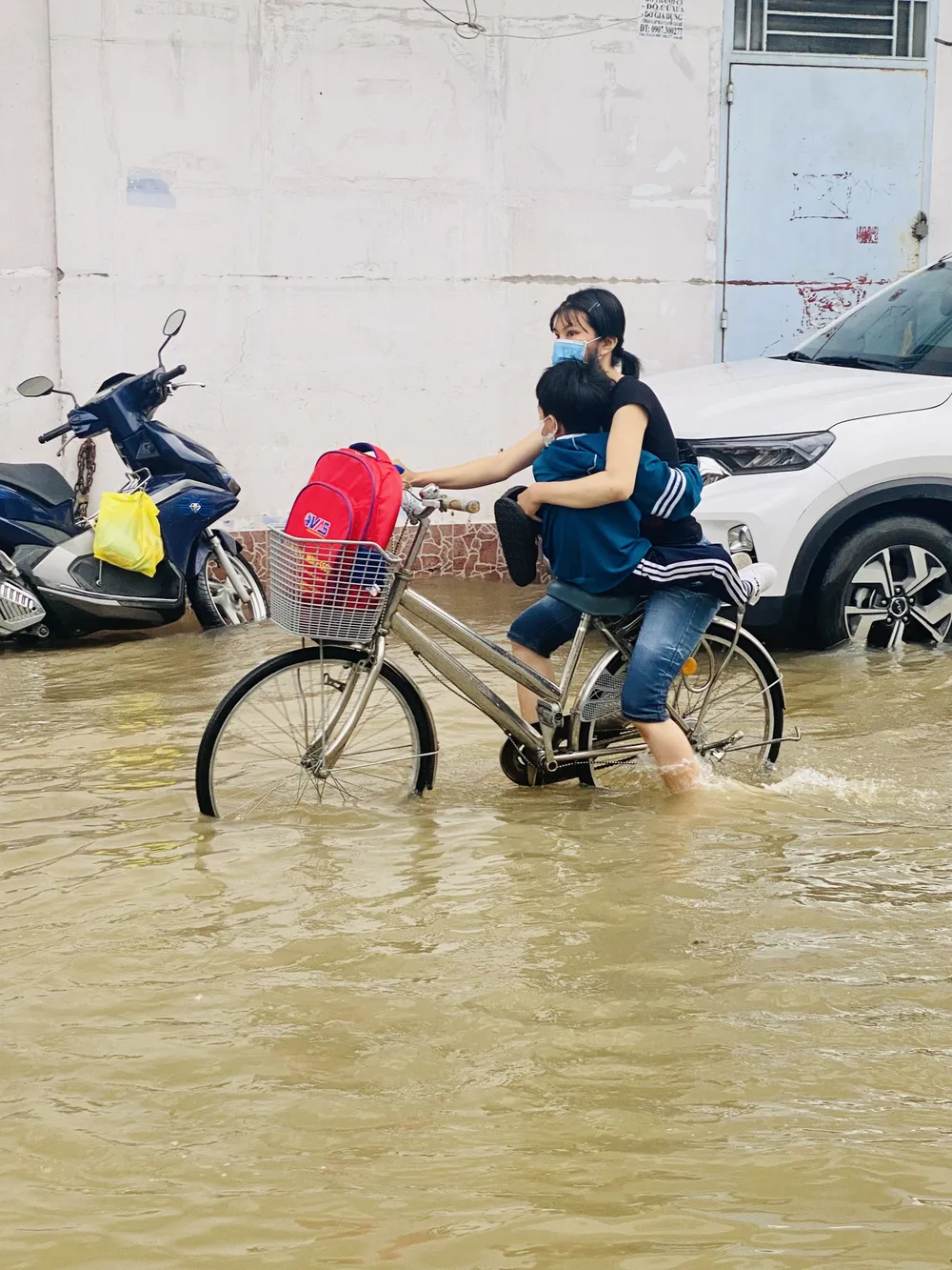 Một cậu bé ôm chặt mẹ trên xe đạp di chuyển qua một con hẻm ngập nặng