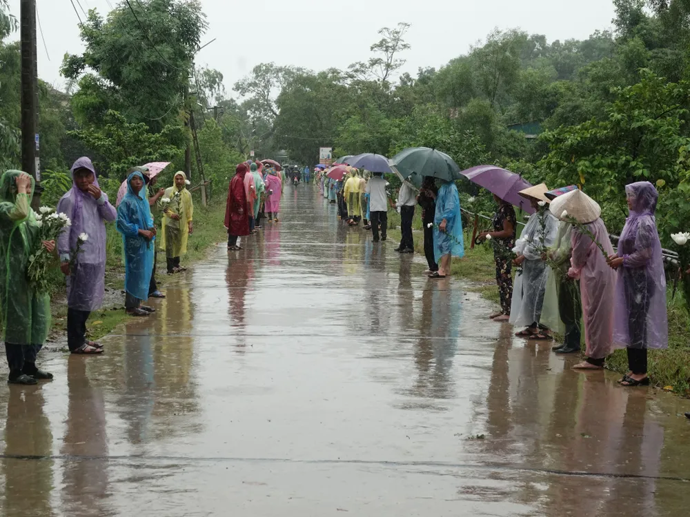 Người dân đội mưa, cầm bông hoa cúc trắng đứng đón linh cữu liệt sĩ Lê Ánh Sáng